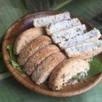 Wooden plate holding coconut-coated ring-shaped bread and several white, coconut-sprinkled slices on a banana leaf background.
