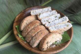 Wooden plate holding coconut-coated ring-shaped bread and several white, coconut-sprinkled slices on a banana leaf background.