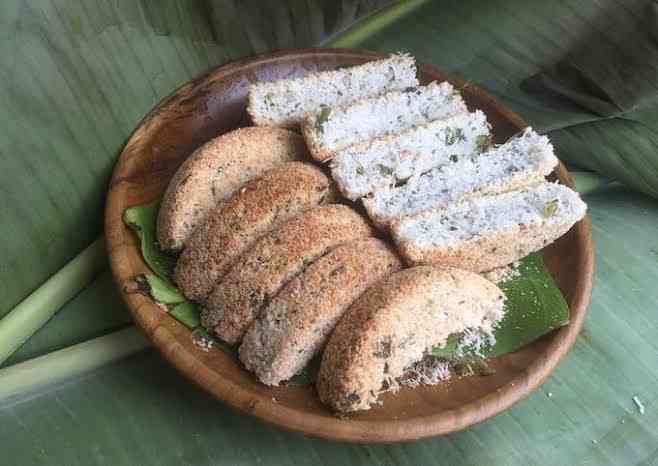 Wooden plate holding coconut-coated ring-shaped bread and several white, coconut-sprinkled slices on a banana leaf background.