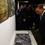 Man in a suit and glasses leans forward to view a black-and-white photo displayed on a gallery table, surrounded by visitors and a photographer in the background.