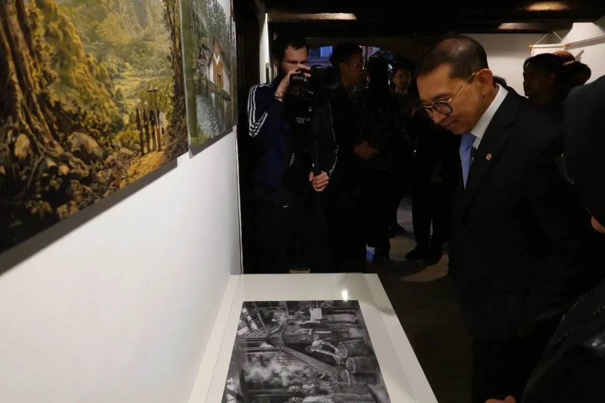 Man in a suit and glasses leans forward to view a black-and-white photo displayed on a gallery table, surrounded by visitors and a photographer in the background.