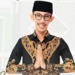 Man in a black batik shirt with gold embroidery, hands in prayer position and smiling at the camera.
