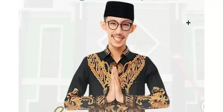 Man in a black batik shirt with gold embroidery, hands in prayer position and smiling at the camera.
