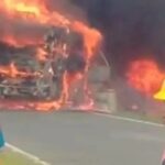 Bus engulfed in flames on a road with firefighters in yellow helmets nearby, smoke billowing around the vehicle.