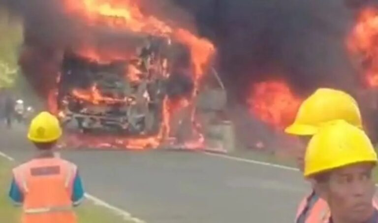 Bus engulfed in flames on a road with firefighters in yellow helmets nearby, smoke billowing around the vehicle.