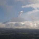 Distant volcano erupting with a plume rising above a layer of clouds over a flat landscape below.