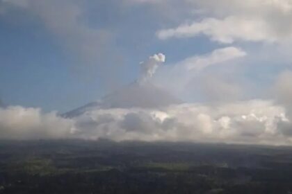 Distant volcano erupting with a plume rising above a layer of clouds over a flat landscape below.