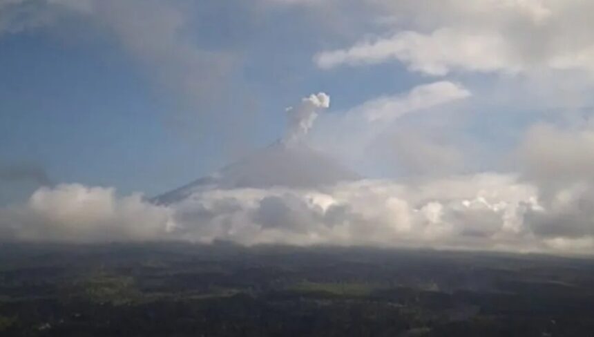 Distant volcano erupting with a plume rising above a layer of clouds over a flat landscape below.