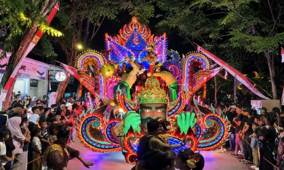 1002781424_1788673755 - Mabur.co Nighttime festival parade with a brightly lit, ornate float featuring a central deity head and dancers, crowds lining the street.
