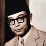 Black-and-white portrait of a man wearing a dark fez, thick-framed glasses, and a light suit, looking toward the camera.