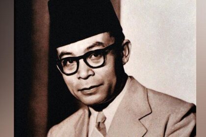Black-and-white portrait of a man wearing a dark fez, thick-framed glasses, and a light suit, looking toward the camera.