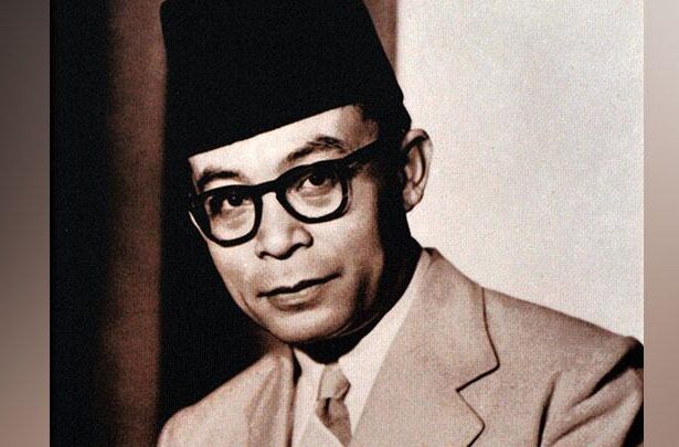 Black-and-white portrait of a man wearing a dark fez, thick-framed glasses, and a light suit, looking toward the camera.