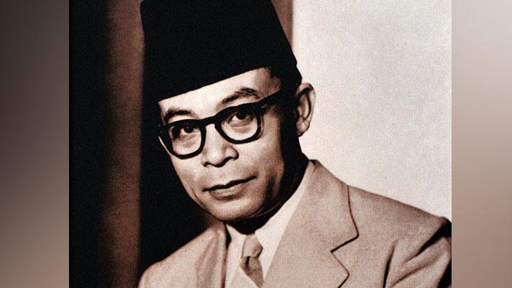 Black-and-white portrait of a man wearing a dark fez, thick-framed glasses, and a light suit, looking toward the camera.
