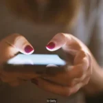 Close-up of hands holding and typing on a smartphone with pink nail polish in low light.