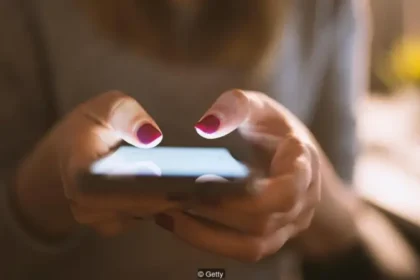Close-up of hands holding and typing on a smartphone with pink nail polish in low light.