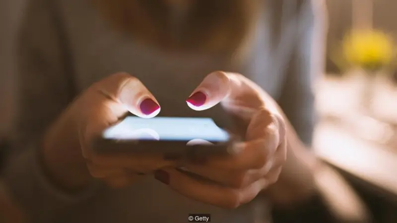 Close-up of hands holding and typing on a smartphone with pink nail polish in low light.