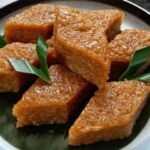 Brown rectangular jaggery-based dessert pieces arranged on a plate with green leaves as garnish, served on a dark tray
