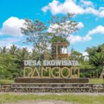Sign reading 'Desa Ekowisata' in an eco-tourism village, with a wooden lookout tower and tropical trees against a blue sky.