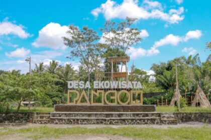 Sign reading 'Desa Ekowisata' in an eco-tourism village, with a wooden lookout tower and tropical trees against a blue sky.