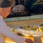 Man in white robes and a black cap arranges gold-wrapped offerings on a wooden floor during a ceremony.