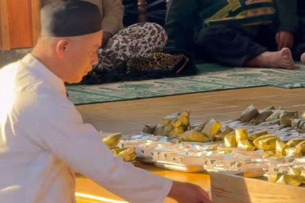 Man in white robes and a black cap arranges gold-wrapped offerings on a wooden floor during a ceremony.