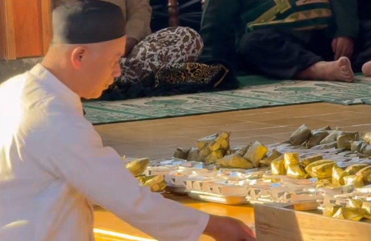 Man in white robes and a black cap arranges gold-wrapped offerings on a wooden floor during a ceremony.