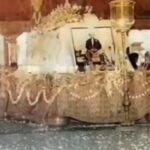 Funeral altar: framed photo of a man on a decorated bier, draped with garlands and beads, mourners nearby.