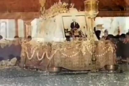 Funeral altar: framed photo of a man on a decorated bier, draped with garlands and beads, mourners nearby.
