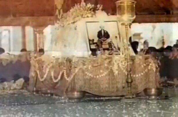 Funeral altar: framed photo of a man on a decorated bier, draped with garlands and beads, mourners nearby.