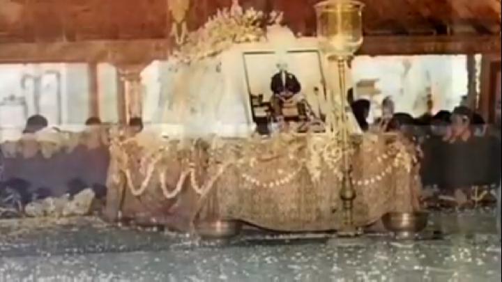 Funeral altar: framed photo of a man on a decorated bier, draped with garlands and beads, mourners nearby.