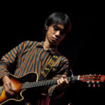 Male guitarist performing on a dark stage, wearing a striped brown and teal shirt while playing a sunburst acoustic guitar with a Fender strap.