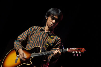 Male guitarist performing on a dark stage, wearing a striped brown and teal shirt while playing a sunburst acoustic guitar with a Fender strap.