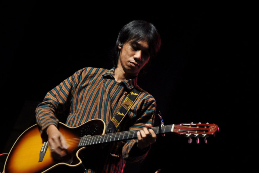 Male guitarist performing on a dark stage, wearing a striped brown and teal shirt while playing a sunburst acoustic guitar with a Fender strap.