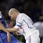 Two soccer players clash for the ball during a match: one in blue on the left and a bald man in white on the right.