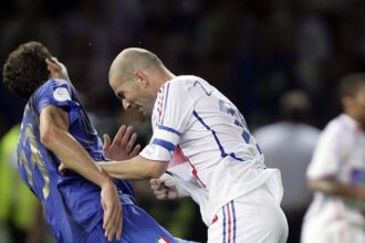 Two soccer players clash for the ball during a match: one in blue on the left and a bald man in white on the right.