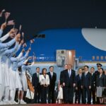President and group of dignitaries pose for a ceremonial welcome on an airport tarmac as supporters wave tiny American flags; airplane in background at night.