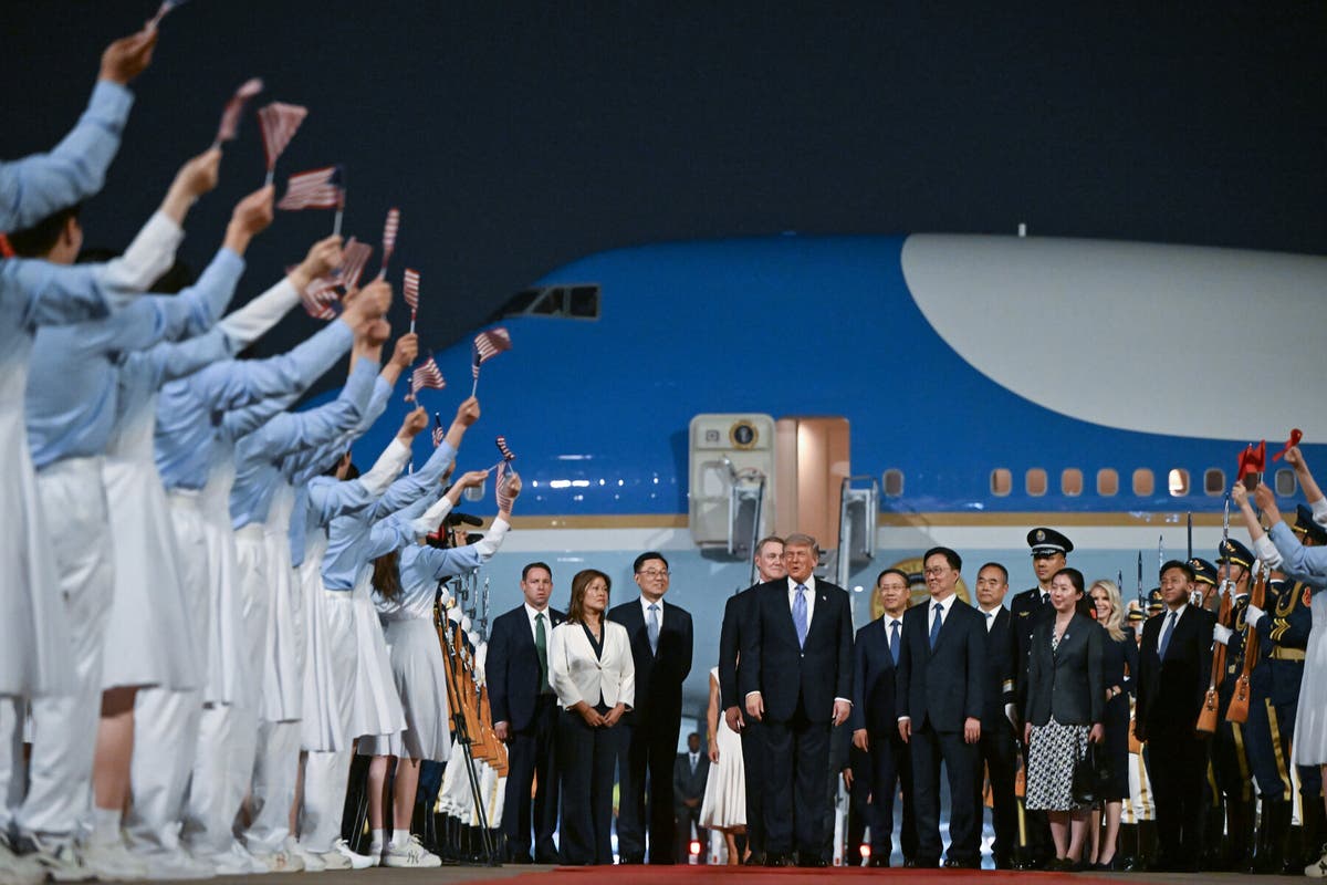 President and group of dignitaries pose for a ceremonial welcome on an airport tarmac as supporters wave tiny American flags; airplane in background at night.