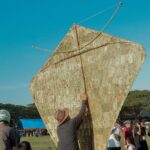 Man raises a giant triangular kite-like sail outdoors at a festival, with a crowd under a clear blue sky.