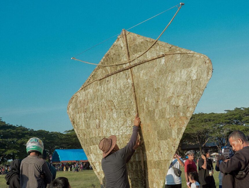 Man raises a giant triangular kite-like sail outdoors at a festival, with a crowd under a clear blue sky.