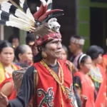 Man in vibrant ceremonial costume with feathered headdress performing at a cultural festival; others in colorful attire watch in the background
