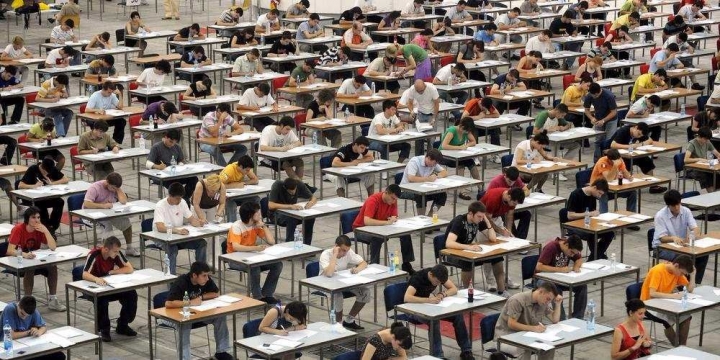 Aerial view of a large exam hall with hundreds of students at individual desks taking tests.