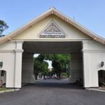 White arch gateway with decorative pediment and tiled crest, opening to a tree-lined campus road beyond.