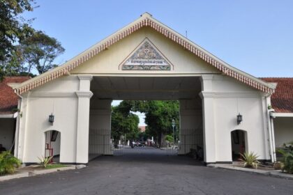 White arch gateway with decorative pediment and tiled crest, opening to a tree-lined campus road beyond.