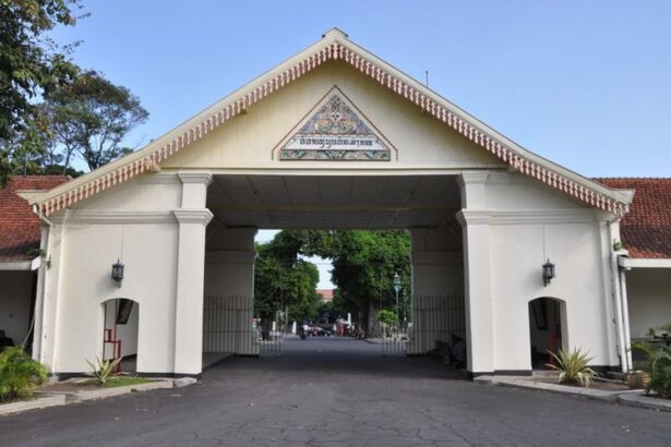 White arch gateway with decorative pediment and tiled crest, opening to a tree-lined campus road beyond.