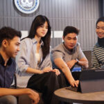 Four young adults gathered around a tablet on a round table, collaborating in a modern lounge.**Note: concise ALT text designed for accessibility.