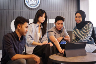 Four young adults gathered around a tablet on a round table, collaborating in a modern lounge.**Note: concise ALT text designed for accessibility.