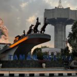 White equestrian statue and a chariot sculpture on a raised platform with a large concrete structure in the background, at Taman Segitiga park.