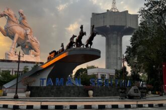 White equestrian statue and a chariot sculpture on a raised platform with a large concrete structure in the background, at Taman Segitiga park.