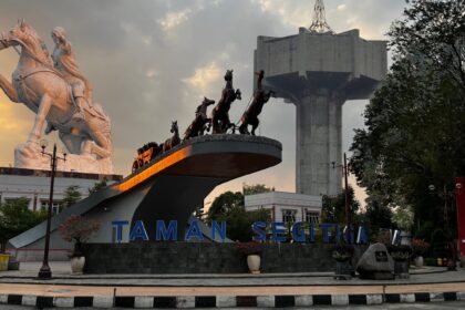 White equestrian statue and a chariot sculpture on a raised platform with a large concrete structure in the background, at Taman Segitiga park.