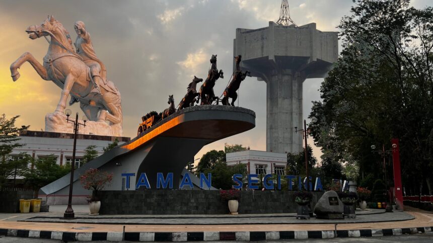White equestrian statue and a chariot sculpture on a raised platform with a large concrete structure in the background, at Taman Segitiga park.